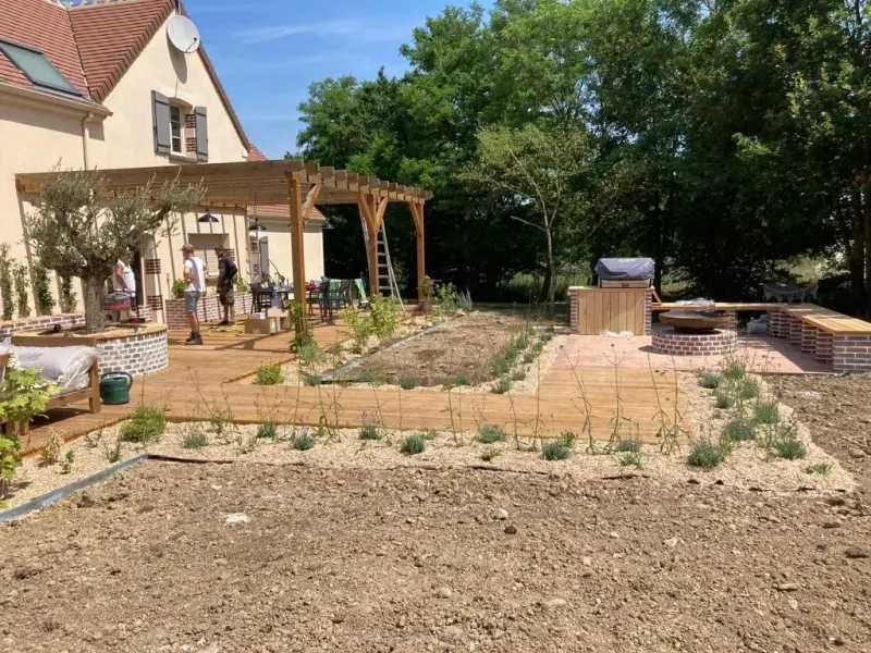 Chantier d'aménagement en cours : terrasse en bois et pergola posées, jeunes plants en godets alignés sur la terre nue avant plantation