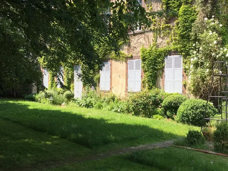 Façade d'une maison de maître aux volets clairs, entièrement couverte de vigne vierge et de rosiers grimpants, devant une pelouse ombragée
