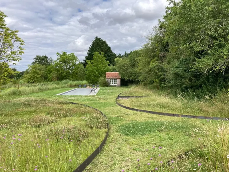 Prairie fleurie fauchée en courbes autour d'un bassin de nage et d'un petit pavillon, en lisière de bois