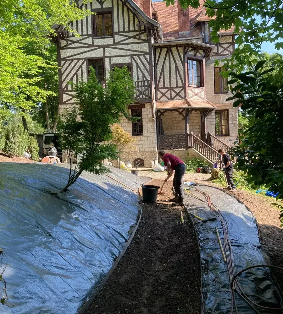 Deux jardiniers plantent un talus couvert de toile de paillage devant une maison à colombages