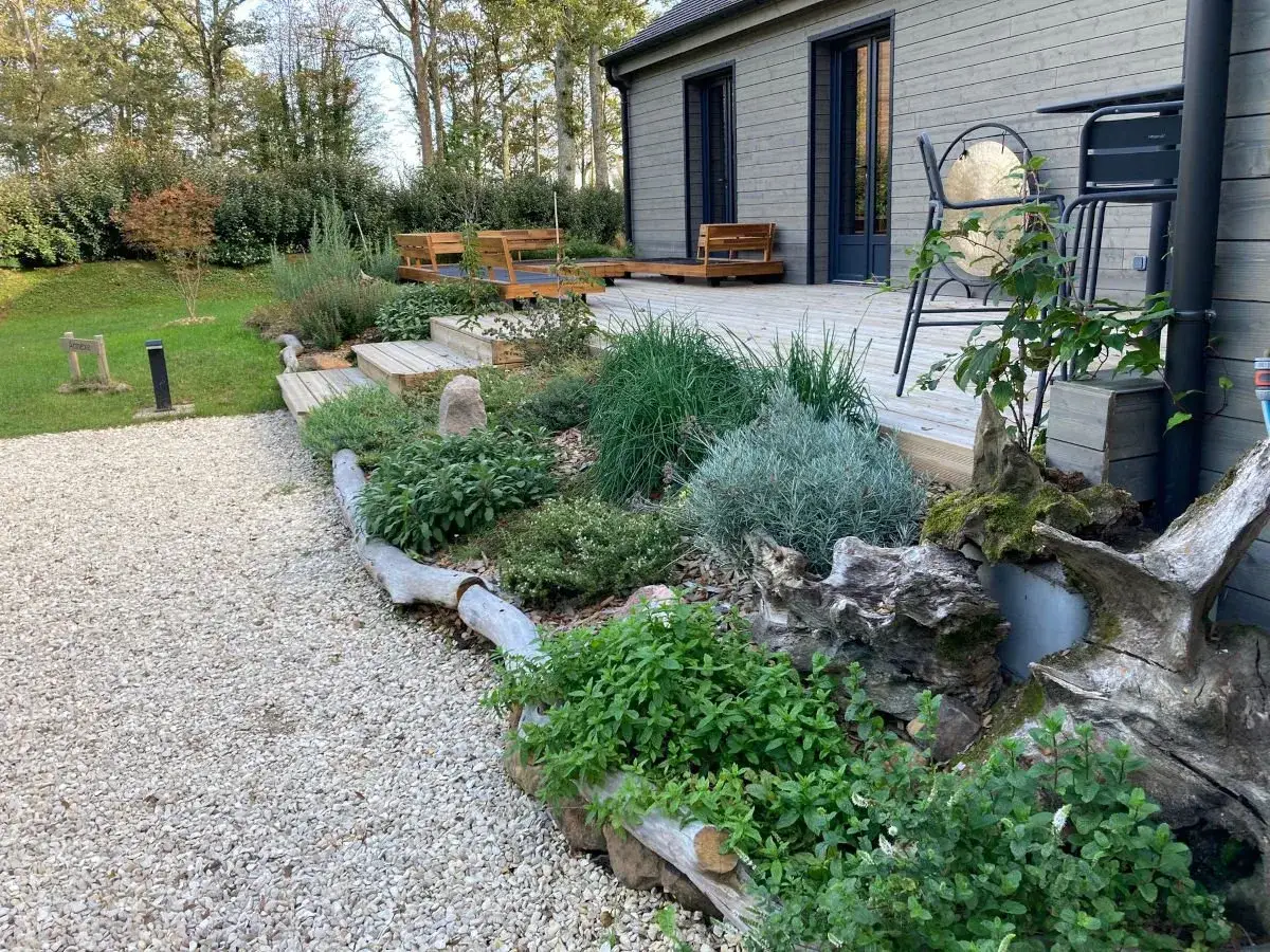 Massif de vivaces et d'aromatiques bordé de bois flotté, entre une allée de gravier clair et une terrasse en bois adossée à une maison au bardage gris