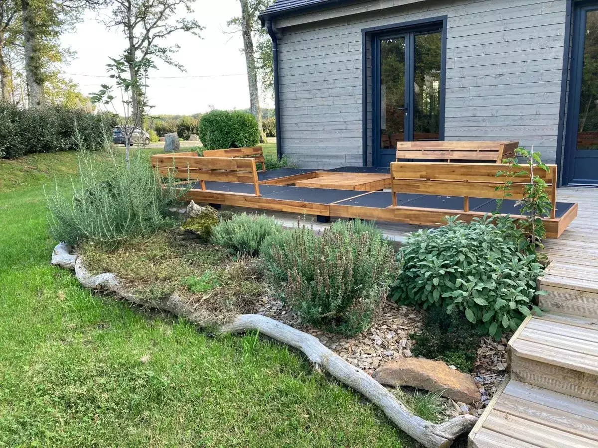 Massif de lavandes, sauges et thyms paillé de copeaux, souligné de troncs flottés, devant une terrasse en bois et ses banquettes