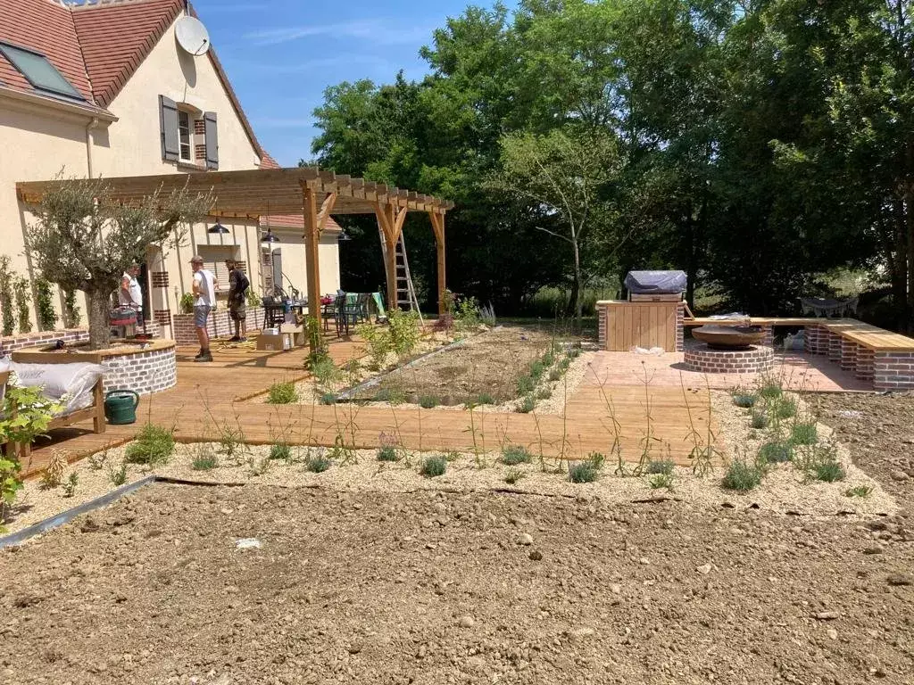 Chantier d'aménagement en cours : terrasse en bois et pergola posées, jeunes plants en godets alignés sur la terre nue avant plantation