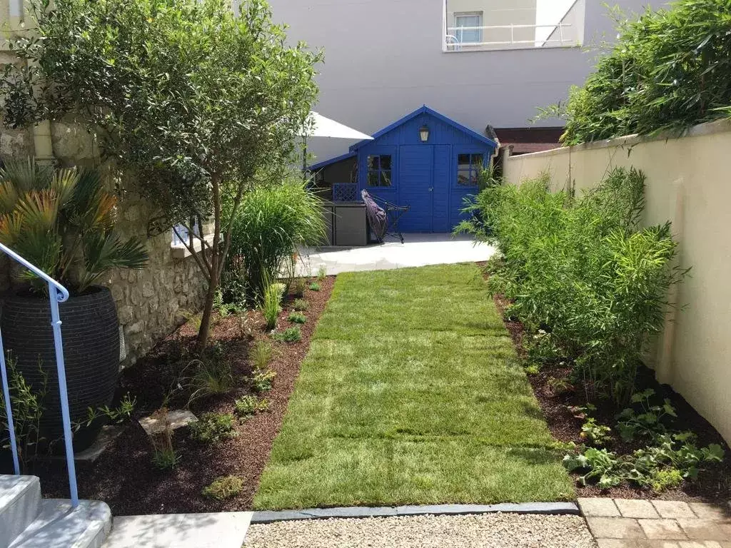 Petit jardin de ville en longueur, bande de gazon fraîchement posée entre deux massifs paillés, cabanon bleu au fond