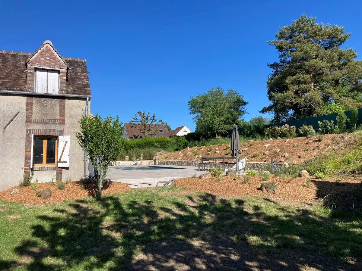 Piscine bordée de rocaille et de massifs paillés, jeune arbre fraîchement planté devant le pignon d'une maison en pierre